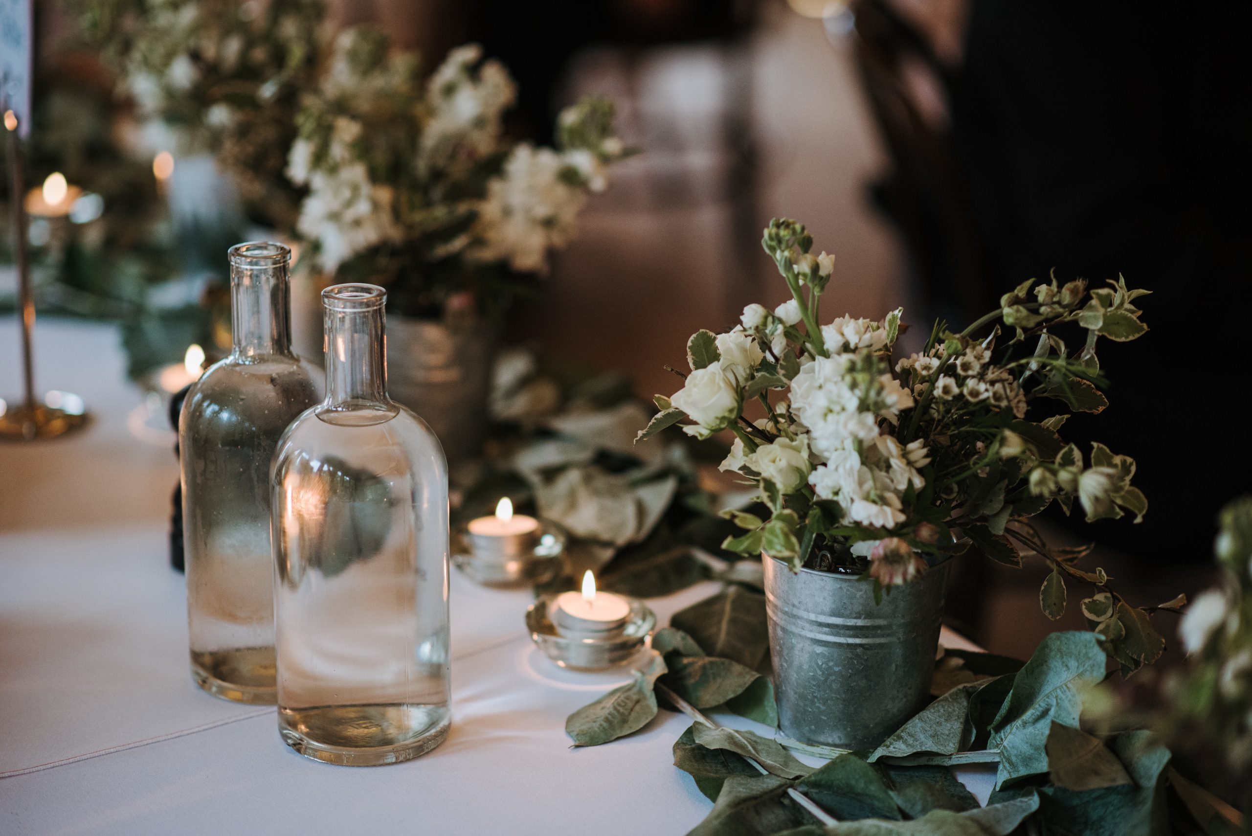 A beautiful shot of white flowers in a bucket, water bottles and candles on a table decorated with leaves